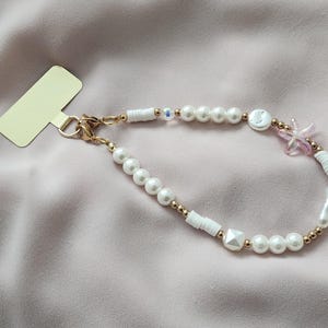 May include: A beaded phone charm with white faux pearls, gold-coloured beads, and white geometric and cylindrical beads. A small pink star-shaped charm and a gold-coloured clasp are also included. The charm is on a pink fabric background.