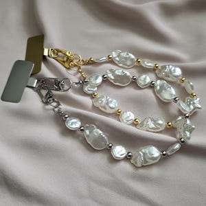May include: A pearl phone strap featuring irregular, white baroque pearls, with small gold and silver beads. The strap has gold and silver metal clasps. The background is a soft, neutral fabric.