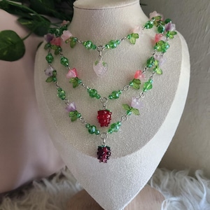 Puede incluir: Un collar de dos hilos con cuentas verdes, rosas y moradas, y cadena plateada. El collar presenta detalles de flores y hojas, con tres colgantes en forma de bayas. El collar se exhibe sobre un busto de maniquí blanco.