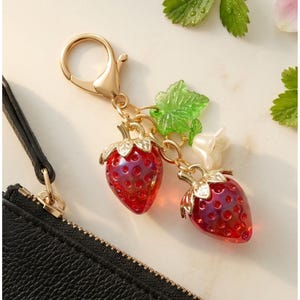 May include: A gold-toned metal keychain featuring two translucent red strawberry charms with textured surfaces, a small green leaf charm, and a white flower charm, all attached to a circular lobster clasp connector.