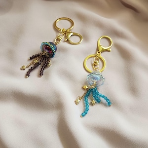 Puede incluir: Dos llaveros de medusas hechos a mano con herrajes dorados. Un llavero presenta una medusa morada y azul con tentáculos de cuentas moradas oscuras. El otro tiene una medusa azul con tentáculos de cuentas turquesas y dijes de estrella dorados.