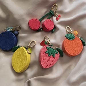 May include: A collection of colorful fruit-shaped keychains. The keychains include a blue circle, a yellow lemon, a pink strawberry, an orange, and a cherry. Each keychain has a gold-colored clasp and a small loop for attaching keys.