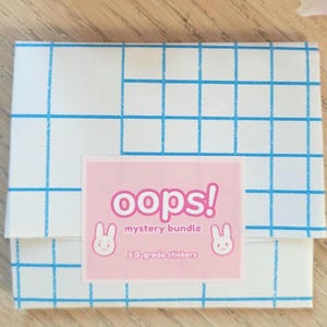 May include: A square white paper envelope featuring a blue grid pattern. A rectangular pink label is centered on the front with the text "oops! mystery bundle" and "5 B-grade stickers" accompanied by two small white rabbit face illustrations.