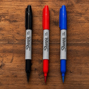 May include: Three Sharpie fine point permanent markers arranged vertically. The markers feature a light gray barrel with the Sharpie logo and text printed in black. The caps and tips are colored black, red, and blue respectively.