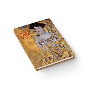 May include: A gold and white journal with a painting of a woman in a gold dress. The painting is a famous work of art by Gustav Klimt.