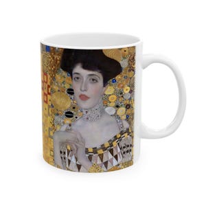 May include: White ceramic mug with a colorful image of a woman with dark hair and a gold background. The woman is wearing a necklace and a patterned dress. The background is filled with gold circles and other geometric shapes.