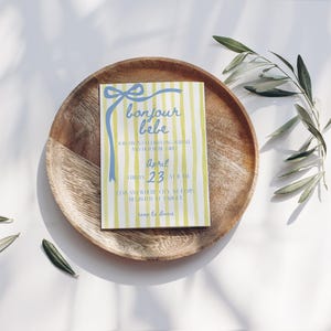 May include: A yellow and blue striped baby shower invitation with a blue bow and the text "bonjour bebe" in blue script. The invitation says "Friday, April 23 at 11 am" and "for an afternoon of love, laughter, and baby shower enjoyment".