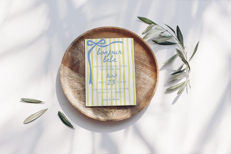 May include: A yellow and blue striped baby shower invitation with a blue bow and the text "bonjour bebe" in blue script. The invitation says "Friday, April 23 at 11 am" and "for an afternoon of love, laughter, and baby shower enjoyment".