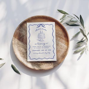 Puede incluir: Una tarjeta de invitación azul y blanca con una cinta azul y un pastel con velas. El texto en la tarjeta dice "Olivia Owen cumple 1 año! Únase a nosotros para celebrar el primer cumpleaños de Olivia. 2 de febrero a las 3 pm. 123 Bagelstreet, Bay City."