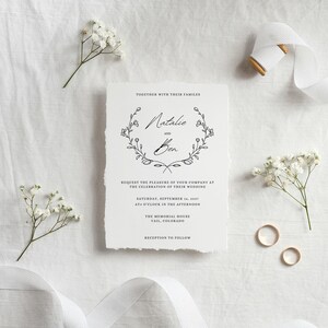 May include: A wedding invitation with a simple floral wreath design. The invitation reads "Natalie and Ben" and includes the date, time, and location of the wedding. The invitation is on a textured, off-white paper.