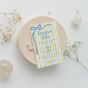 May include: A yellow and blue striped baby shower invitation with the text "bonjour bebe" and details for the event. The invitation is on a light pink surface.