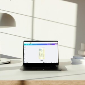 May include: A laptop computer with a white background is open to a design program. The screen shows a yellow and white design with a blue ribbon. The design is for a gift tag.