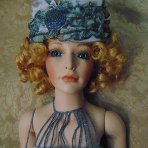 May include: A porcelain doll with blonde curly hair, blue eyes, and a pale complexion. The doll wears a light blue beaded halter top and a matching hat with a large bow and beaded embellishments. The doll's lips are a soft coral color.