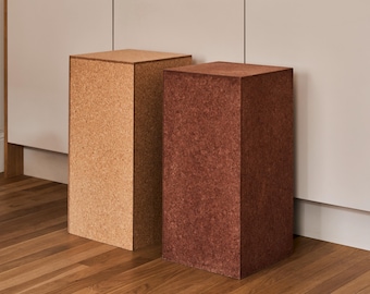 May include: Two square-shaped cork pedestals, one is light brown and the other is a darker reddish brown. The pedestals are standing on a wooden floor.