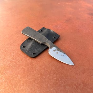 Puede incluir: Un pequeño cuchillo forjado a mano con una hoja puntiaguda y un mango oscuro y texturizado. El cuchillo descansa sobre una funda de cuero negro. La hoja muestra signos de desgaste, lo que aumenta su atractivo rústico. El diseño general sugiere un enfoque en la funcionalidad y la artesanía.
