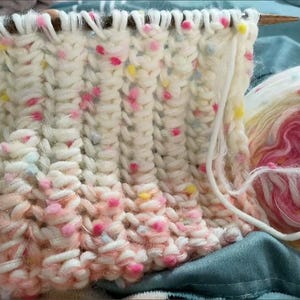Könnte beinhalten: Nahaufnahme eines gestrickten Stoffes mit weißer Basis und bunten Flecken in Rosa, Blau und Gelb. Der Stoff wird mit Stricknadeln und Garn bearbeitet.