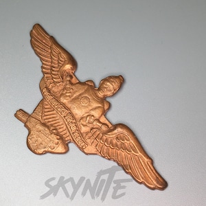 Puede incluir: Un pin de metal de color dorado con una figura alada que sostiene una espada y un escudo. El pin tiene el texto "Buck Rogers, Solar Scouts" inscrito en él.