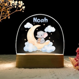 May include: A night light featuring a cartoon elephant holding a teddy bear, sitting on a crescent moon. The name "Noah" is written above the moon. The light sits on a wooden base and is surrounded by clouds and stars.