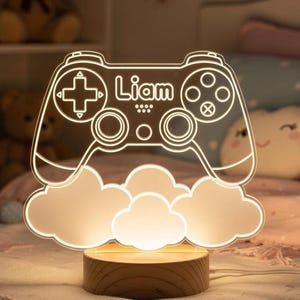 Könnte beinhalten: Nachtlicht im Gaming-Design mit Controller-Form und dem Namen "Liam" in Weiß. Das Design umfasst Wolkenformen und steht auf einem Holzsockel. Ideal für ein Kinderzimmer.