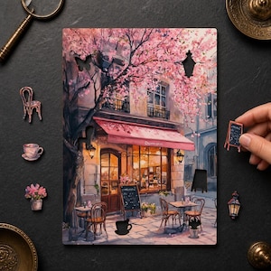 Könnte beinhalten: Ein rechteckiges Holzpuzzle-Brett mit einer gedruckten Illustration eines Cafés im Pariser Stil mit einem rosa Kirschblütenbaum, einer rosa Markise und Sitzgelegenheiten im Freien. Mehrere kleine, separate Puzzleteile aus Holz sind enthalten.