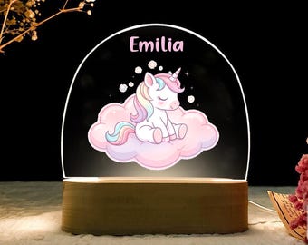May include: A night light featuring a cartoon unicorn sitting on a pink cloud. The unicorn has a rainbow mane and horn. The name "Emilia" is printed above the unicorn. The light sits on a wooden base.