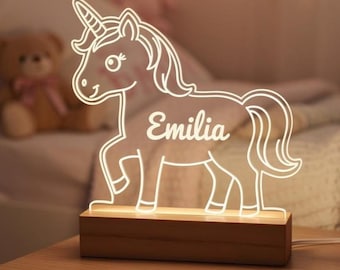 May include: A glowing unicorn-shaped night light with the name "Emilia" etched on its side. The light is made of clear acrylic and sits on a rectangular wooden base. The unicorn has a horn, mane, and tail.