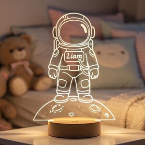 Op de afbeelding: Een decoratief nachtlampje met een astronautenontwerp. De astronaut is geëtst in een helder acryl paneel, verlicht door een warm licht van een houten voet. De naam "Liam" is zichtbaar op de borst van de astronaut.