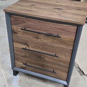 May include: A three-drawer dresser with a dark wood finish and black metal hardware. The dresser has a gray painted frame and sits on four black metal legs.