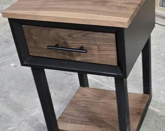 May include: A small wooden end table with a black metal frame. The table has a single drawer and a lower shelf. The drawer has a black metal handle.