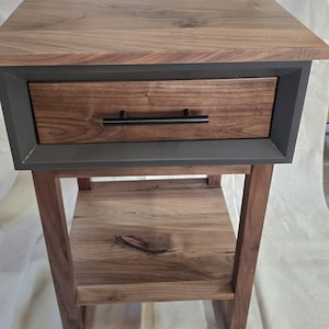 Puede incluir: Una mesa auxiliar de madera con un cajón y un estante inferior. La mesa está hecha de madera de nogal y tiene un borde pintado de gris oscuro alrededor del cajón. El cajón tiene una manija de metal negro.
