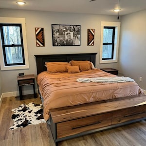 May include: A bedroom with a large bed with a brown headboard and a wooden footboard with storage drawers. The bed has a brown and orange comforter and a white throw blanket. There are two windows on either side of the bed, and a nightstand with a lamp on the right side of the bed.