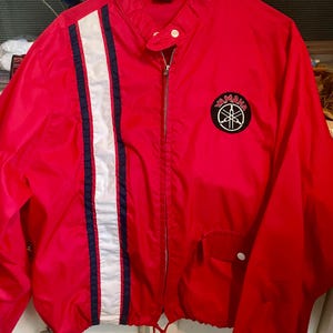May include: A red Yamaha jacket with a white and navy stripe down the left side. The jacket has a zipper closure, a small pocket, and a Yamaha logo patch on the chest. The jacket has a drawstring at the bottom.