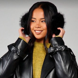 May include: Black faux fur earmuffs with a soft, fluffy texture. The earmuffs are worn by a person wearing a black leather jacket and a yellow sweater. The earmuffs are designed to keep ears warm in cold weather.