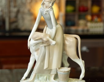 May include: A porcelain figurine depicting a woman in a long dress and head covering, tending to a cow. The figurine is primarily cream-colored with light blue and tan accents. A small bucket sits at the base.