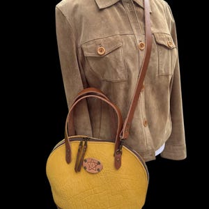 May include: A mustard yellow handbag with a brown leather base and handles. The bag has a textured surface and a brown leather strap. A brown suede jacket is in the background.