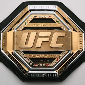 Pu&ograve; includere: Una cintura da campionato con cinturino in pelle nera e una grande placca centrale color oro. La placca presenta le lettere "UFC" in grassetto, circondate da accenti argentati e dorati e piccole pietre preziose.