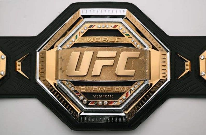 Pu&ograve; includere: Una cintura da campionato con cinturino in pelle nera e una grande placca centrale color oro. La placca presenta le lettere "UFC" in grassetto, circondate da accenti argentati e dorati e piccole pietre preziose.