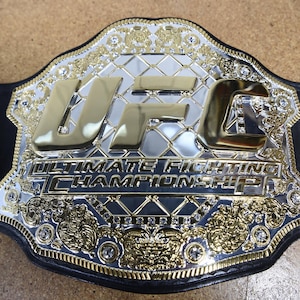 Pu&ograve; includere: Primo piano di una cintura da campionato UFC. La cintura presenta una grande placca centrale argentata con le lettere dorate "UFC" e le parole "Ultimate Fighting Championship". La cintura ha un cinturino in pelle nera e dettagli dorati.