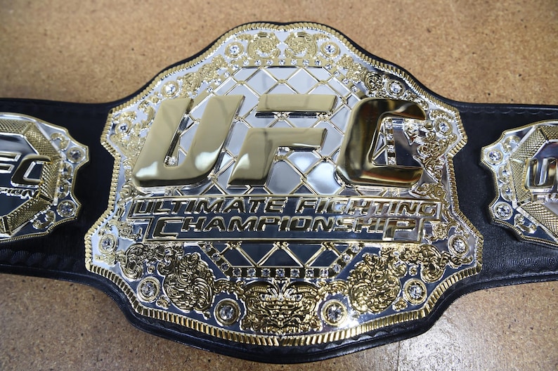 Pu&ograve; includere: Primo piano di una cintura da campionato UFC. La cintura presenta una grande placca centrale argentata con le lettere dorate "UFC" e le parole "Ultimate Fighting Championship". La cintura ha un cinturino in pelle nera e dettagli dorati.
