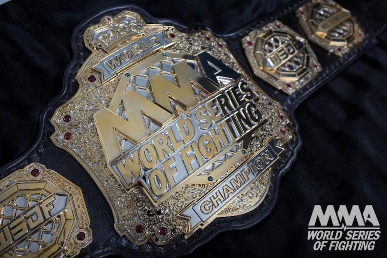 Pu&ograve; includere: Una cintura da campionato MMA World Series of Fighting in oro e argento. La cintura presenta le parole "MMA World Series of Fighting Champion" e il logo MMA. La cintura &egrave; su uno sfondo nero.