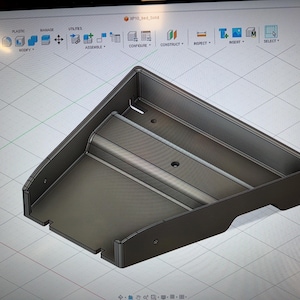 May include: A dark gray, angular, rectangular container with a sloped top and a flat base. The design features a central raised bar and several small circular holes. The object is displayed on a grid background.