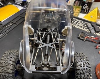 May include: A clear plastic RC car body with a black chassis and suspension components. The car has large, knobby tires and visible internal mechanisms. The body has a streamlined design with a transparent windshield.