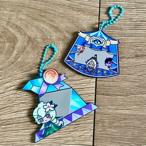 Pode incluir: Dois chaveiros de acrílico com correntes de contas azuis. Um é um design de chapéu de bruxa azul e roxo com um personagem de desenho animado. O outro é um design de tenda azul com personagens de desenhos animados. Ambos os chaveiros têm um acabamento brilhante e iridescente.