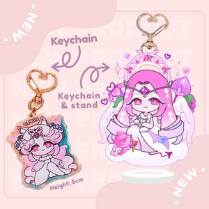 May include: Two acrylic keychains with cartoon character designs. One keychain features a pink character with a crown and the text "Height: 5cm". The other keychain has a pink character with wings. Both keychains have gold-colored heart-shaped clasps.