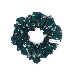 May include: A dark green scrunchie featuring a holly and berry pattern with white dots. The scrunchie has a label that reads "tied up & knotty". A festive hair accessory.