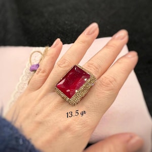 May include: A large, rectangular ruby-red gemstone ring with a gold-colored band. The band is encrusted with small, sparkling stones. The ring is displayed on a hand. The text "13.5 gr" is visible on the image.