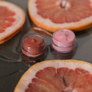 Könnte beinhalten: Zwei kleine Glasbehälter mit Deckeln, die rosa und braune Cremes enthalten, auf einer grauen Oberfläche mit zwei Grapefruit-Hälften.