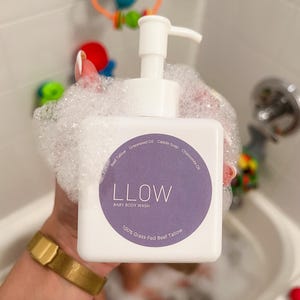 Könnte beinhalten: Weiße Baby-Körperwaschflasche mit Pumpspender, umgeben von Blasen. Das Etikett ist lila mit dem Markennamen "LLOW" und "BABY BODY WASH". Zutaten sind Weidefett. Eine goldene Uhr ist sichtbar.