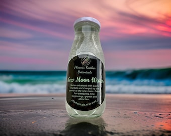 May include: A clear glass bottle with a black label that reads "New Moon Water" and "Phoenix Feather Botanicals". The bottle is sitting on a beach with a sunset in the background.