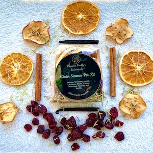 May include: A close-up of a Winter Simmer Pot Kit from Phoenix Feather Botanicals. The kit includes dried orange slices, apple slices, cranberries, cinnamon sticks, cloves, rosemary, nutmeg, and vanilla beans. The kit is packaged in a clear plastic bag with a black label that says "Winter Simmer Pot Kit".
